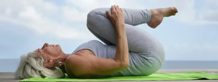 Older woman lying on yoga mat, leg bent, pulling knees toward chest